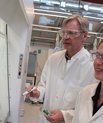 [Translate to English:] Nina Lock og Troels Skrydstrup iført hvide kitler og beskyttelsesbriller tegner formler på glas i laboratorium.