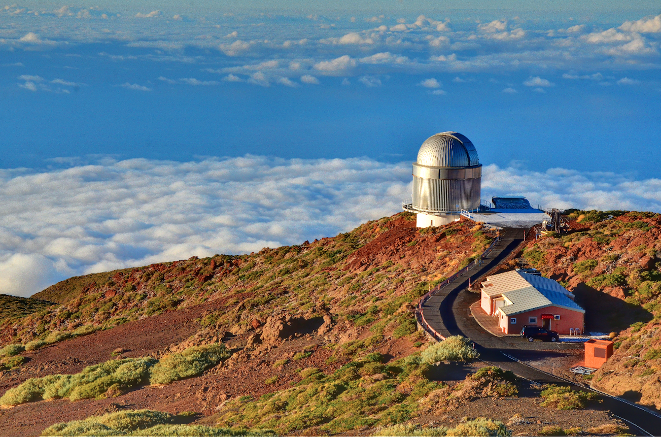 Observatoriet med den sølvskinnende kuppel på en bjergtop over skyerne i baggrunden, med en servicebygning ved siden af.